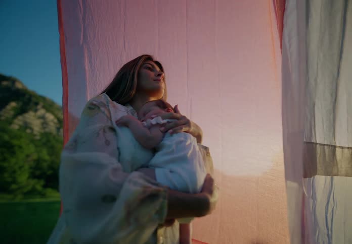 Mother in a dress holding a newborn baby girl while standing in a field with mountains in the background