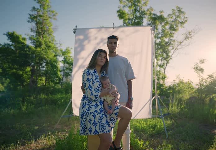 A couple with a newborn baby standing in front of a white photography backdrop outdoors