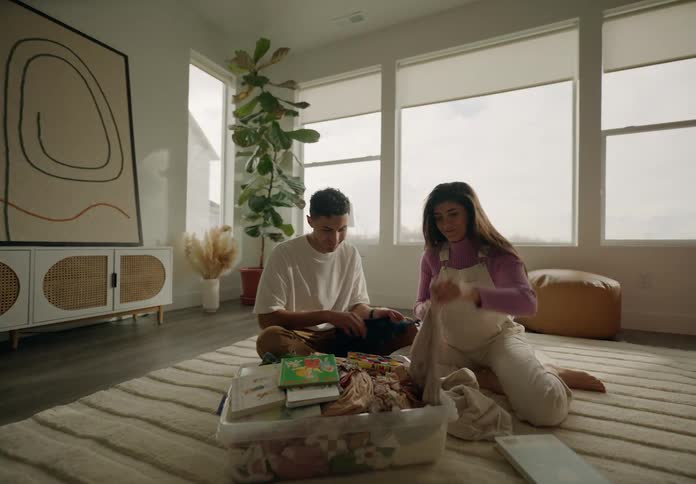 A couple sitting on the floor folding laundry from a basket in a welllit cozy living room with plants and furniture
