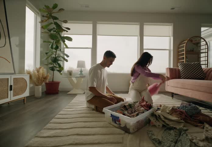 A couple sitting on the floor folding laundry from a basket in a well lit cozy living room with plants and furniture