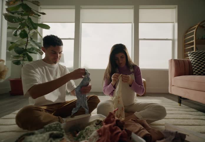 A couple sitting on the floor folding laundry from a basket in a welllit cozy living room with plants and furniture
