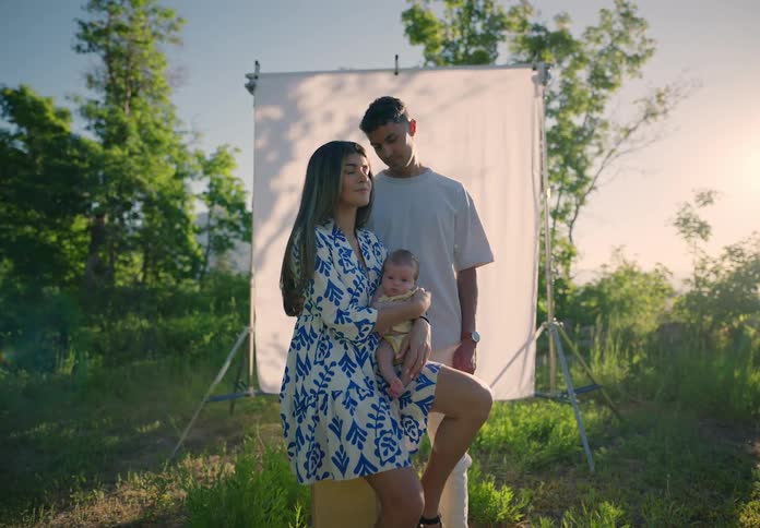 A young couple stand with a baby in front of a white backdrop outdoors at sunset or sunrise