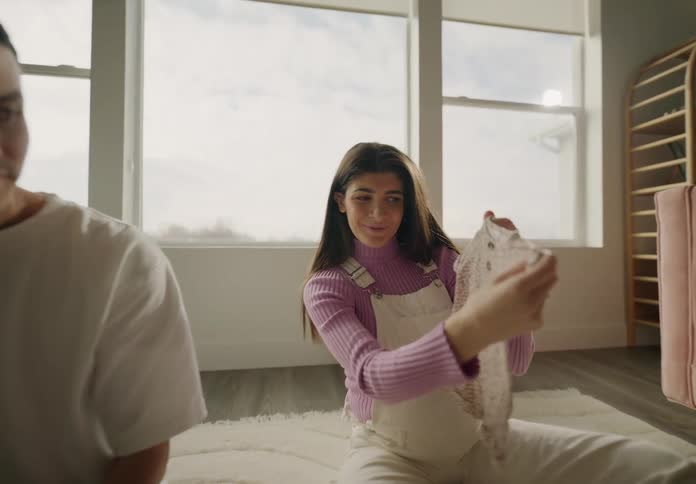 A couple sitting on the floor folding laundry from a basket in a welllit cozy living room with plants and furniture