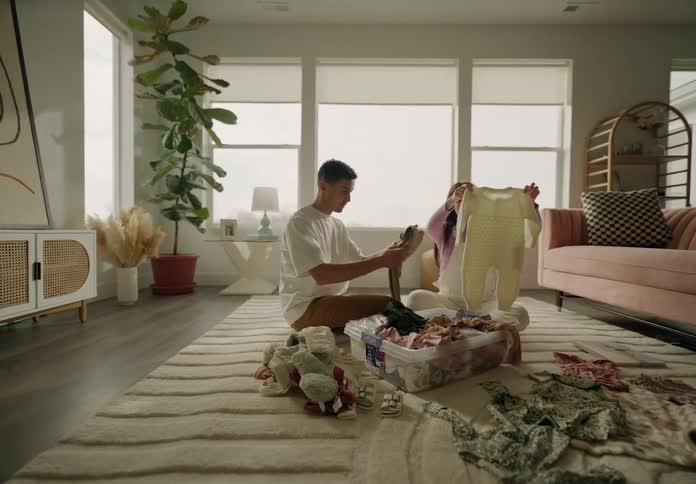 A couple sitting on the floor folding laundry from a basket in a welllit cozy living room with plants and furniture