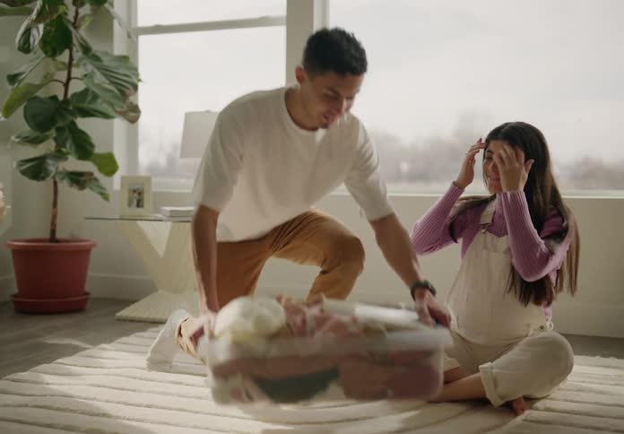 A couple sitting on the floor folding laundry from a basket in a welllit cozy living room with plants and furniture