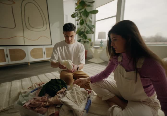 A couple of a man and a woman sitting in a living room fiddling with a coloring book and baby clothes and the woman is pregnant