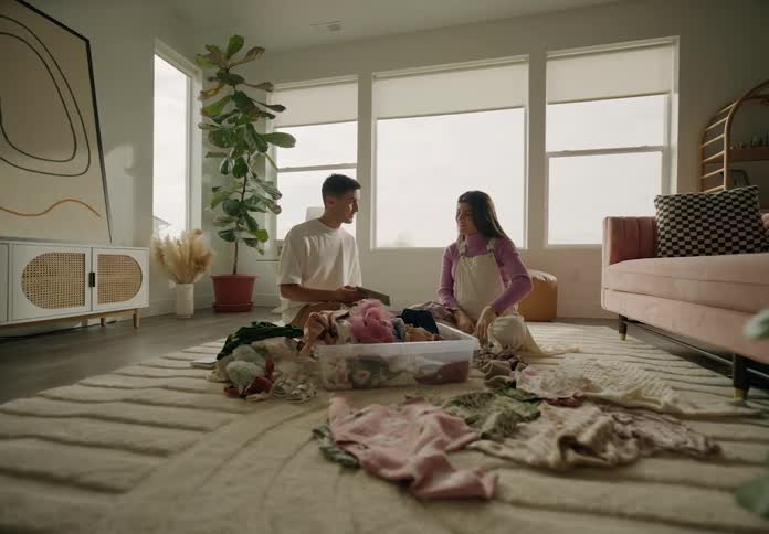 A couple sitting on the floor folding laundry from a basket in a welllit cozy living room with plants and furniture