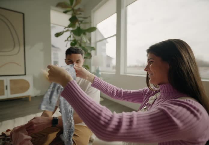 A young couple sits in the living room admiring a baby onesie