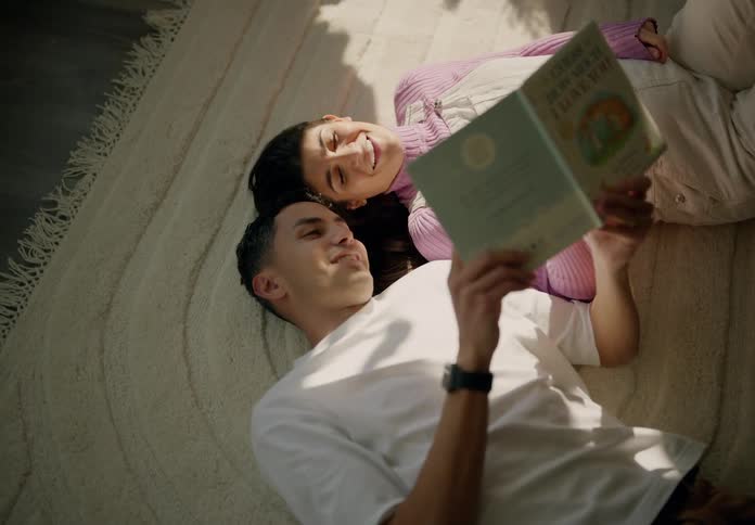 A couple lies on the bed their heads resting against each other reading a book in the morning