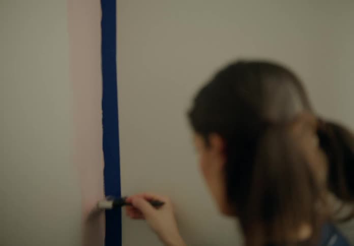 A person painting a wall using a brush to apply a pink color next to a blue tape line
