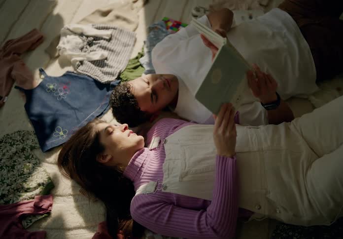 Man and woman lying together on the carpet reading a book and baby clothes in the background
