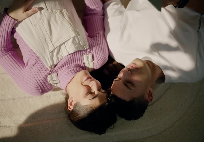 A couple lies on the bed their heads resting against each other reading a book in the morning