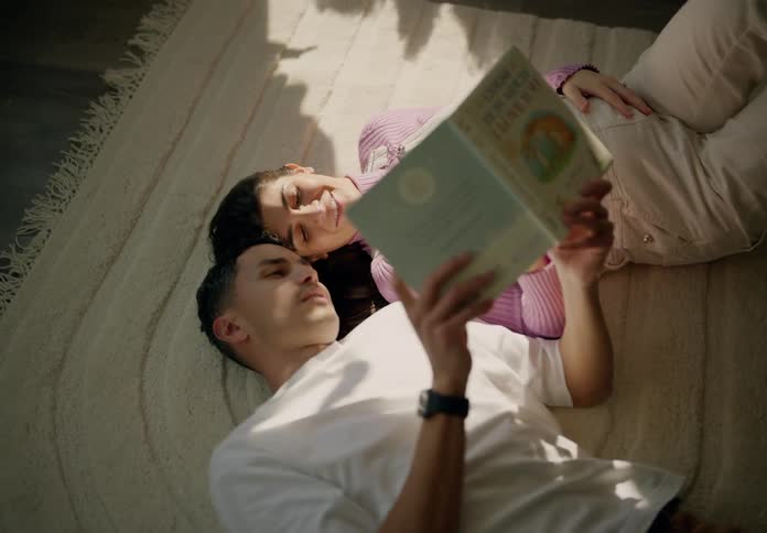 A couple lies on the bed their heads resting against each other reading a book in the morning