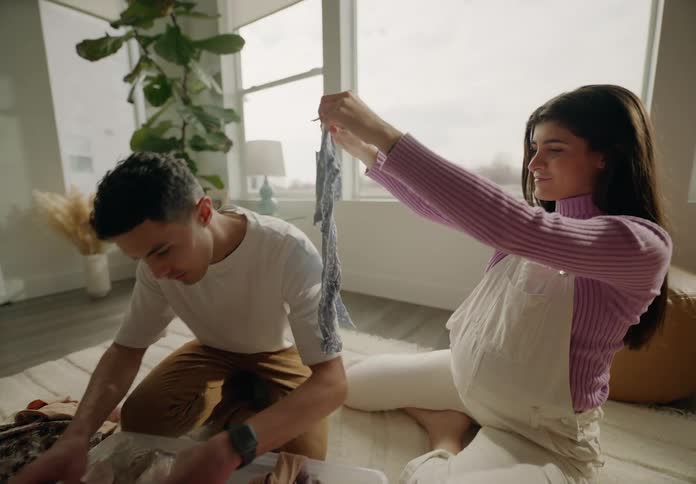 A man and a woman sitting are taking baby clothes from a plastic basin and are happy
