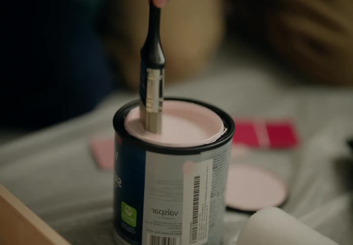 A closeup of a hand dipping a paintbrush into an open can of pink paint
