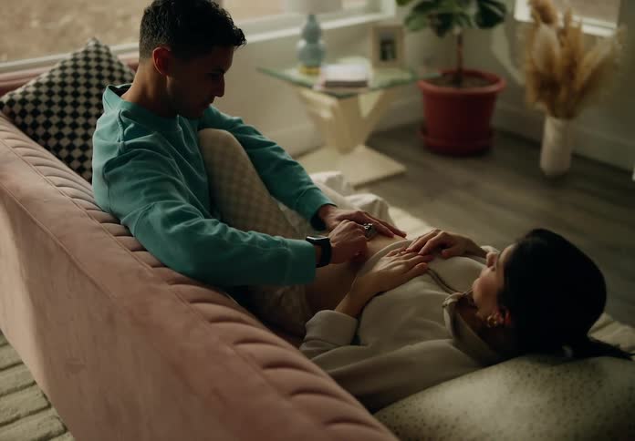 Man sitting on the couch next to his pregnant wife lying down and playing with a crank on her belly