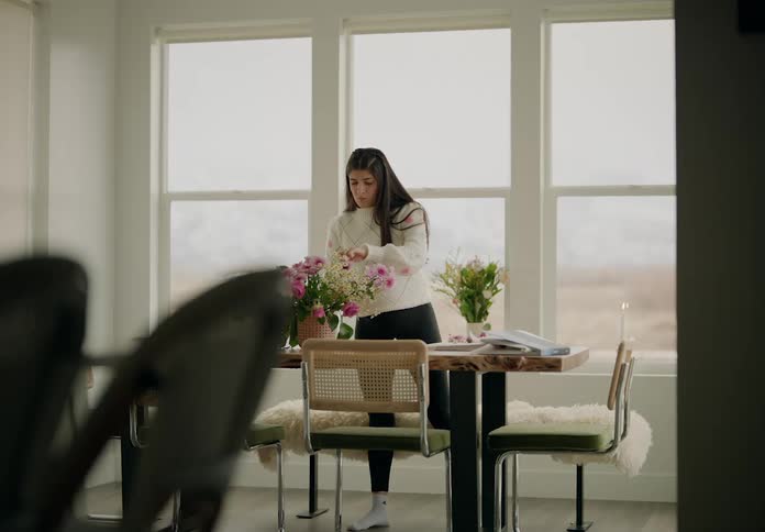 A woman stands next to large windows arranging a bouquet of pink and white flowers