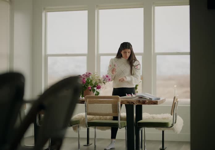 A woman in a white coat is standing in front of a table with a book and a vase of flowers