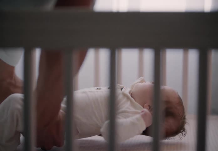 A man is looking at his son who is sleeping on his lap and he puts him in a crib