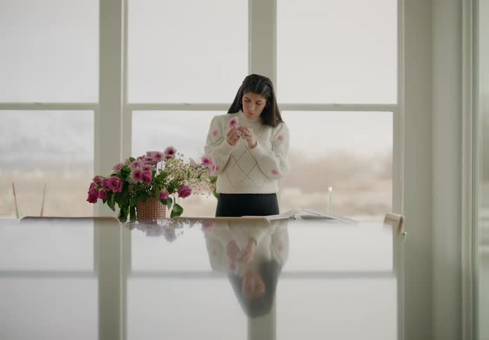 A woman stands next to large windows arranging a bouquet of pink and white flowers