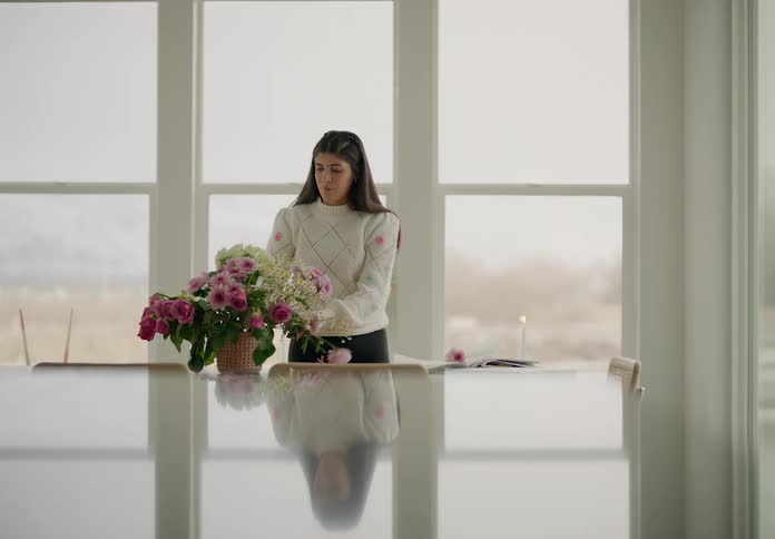 A woman with long brown hair is in a room arranging flowers in a pot