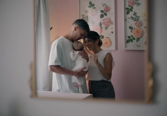 Man standing holding a baby in his arms and giving his wife a kiss seen through a mirror