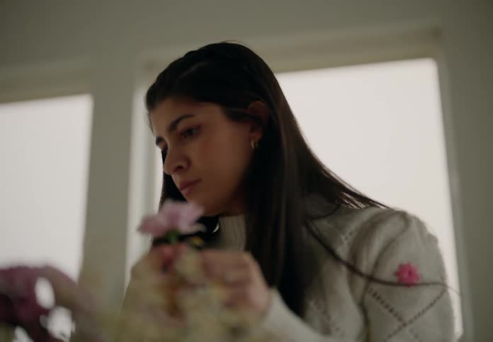 A woman stands next to large windows arranging a bouquet of pink and white flowers