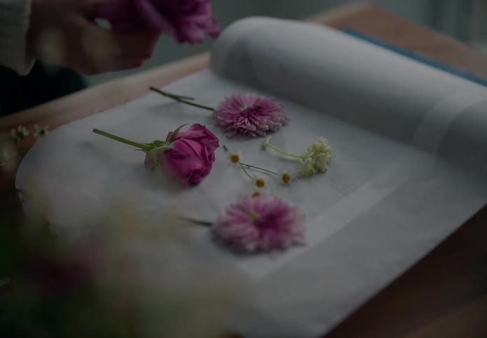 A persons hands carefully arranging pink roses and other flowers on a sheet of parchment paper