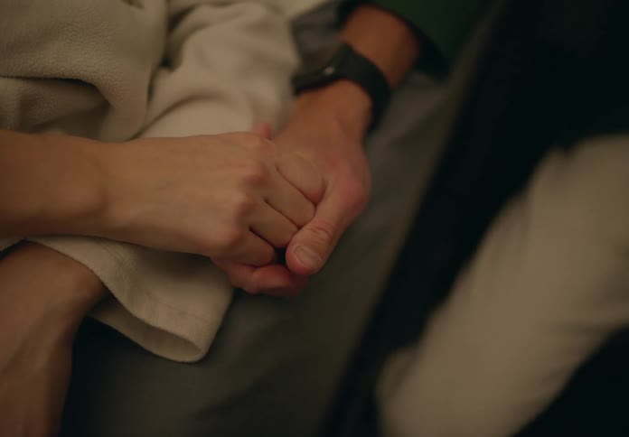 A closeup of two people holding hands one on a bed the other sitting beside