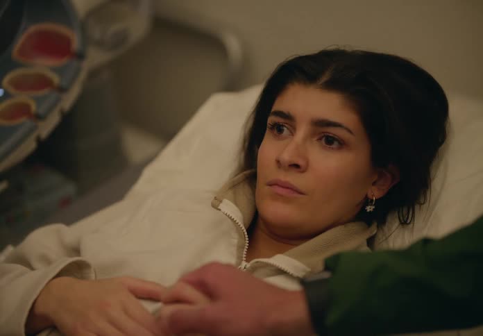 A pregnant young woman in a bed next to an ultrasound machine and holding a mans hand smiles as she talks to someone