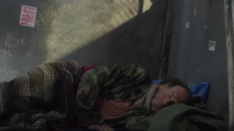 A homeless man lies down against a wall to sleep