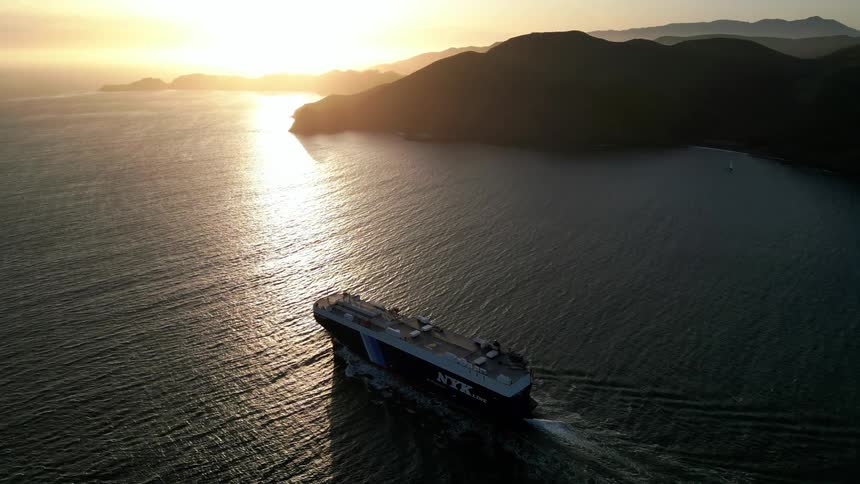 Aerial view of a large ship traveling at sea with the coast in the background and the sun on the horizon