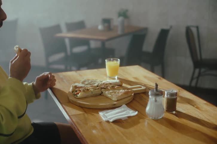 A person is eating sitting at a wooden table with a plate of food orange juice in a glass and condiment shakers