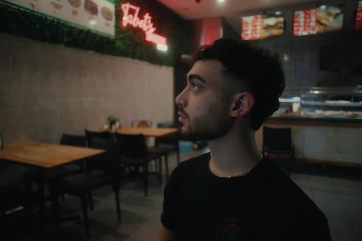 A young man looking around and smiling in a dimly lit restaurant with wooden tables and a neon sign in the background