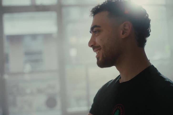 A young man sits at a cafe table talking to a friend while sunlight filters through large windows