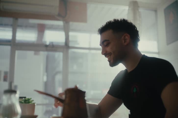 A young man sits at a cafe table sipping coffee while sunlight filters through large windows