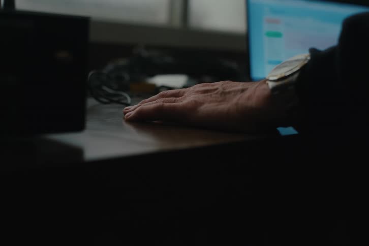 An arm rests on a desk with a computer monitor displaying colorful graphs or charts in the background