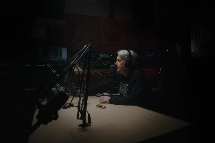 A radio host wearing headphones sits in a booth and speaks into a microphone inside a dimly lit broadcasting studio