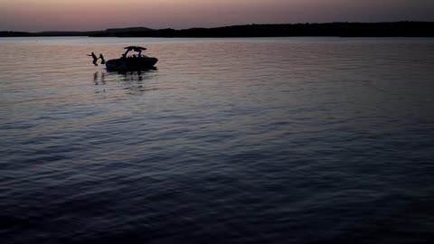 Two people are jumping off a boat into a calm lake in slow motion at dusk