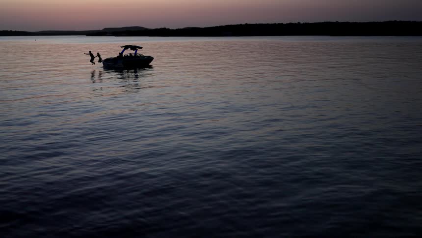 Two people are jumping off a boat into a calm lake in slow motion at dusk