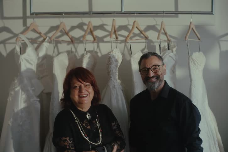 Two people smile in front of the wedding dresses