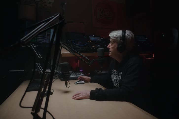 A person sitting at a radio station desk wearing headphones and speaking into a microphone with dj equipment visible in the background