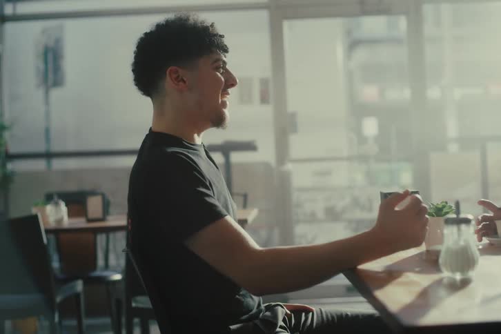 A young man sits at a cafe table talking to a friend while sunlight filters through large windows