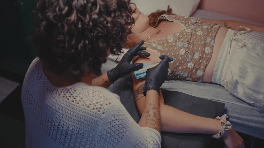 A woman wearing a black gloves is tattooing the arm of a woman who is lying down