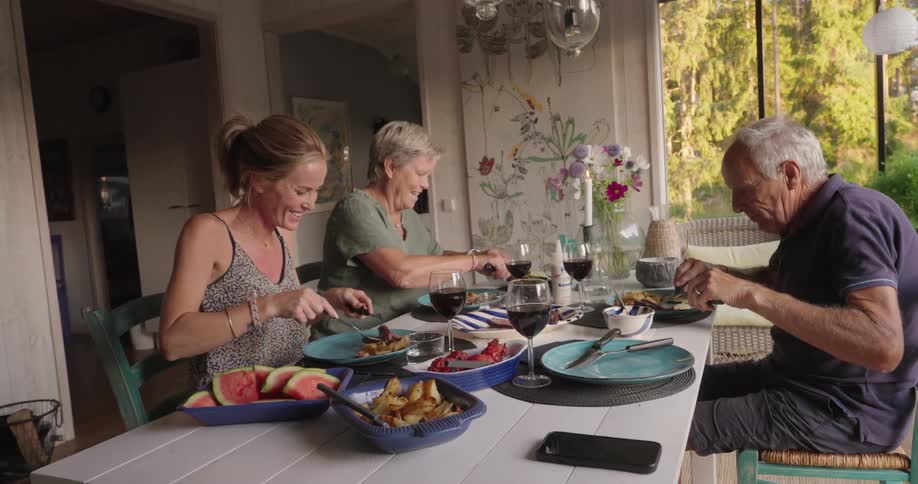 A family sits around a dining table enjoying a meal in a room with large windows overlooking a forest
