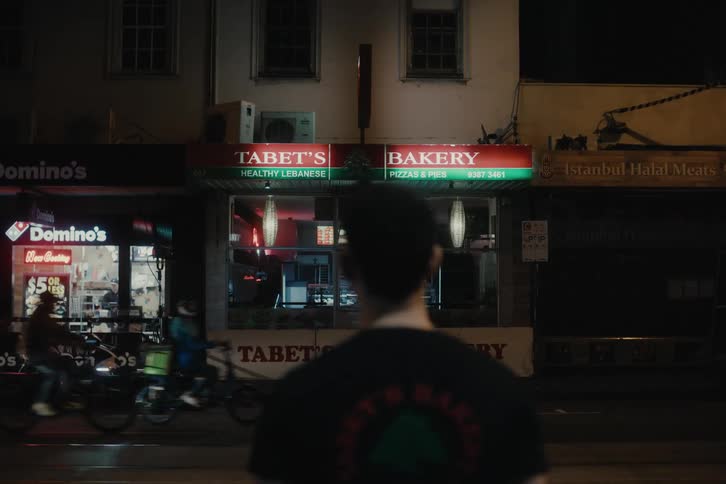 Man wearing t shirt with a bakery logo stands across a busy street from the bakery at night