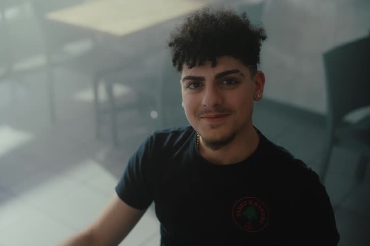 A young man is wearing a partial smile as he looks straight ahead and behind him are some tables and chairs