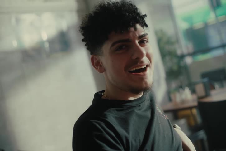A young man sits at a cafe table talking to a friend while sunlight filters through large windows