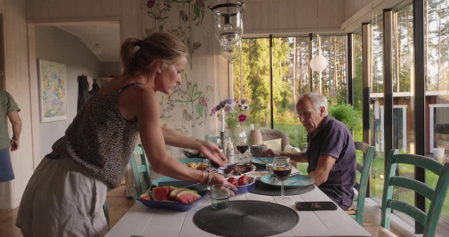 A woman placing a tray of food on a table with glasses and plates and there is a gentleman sitting at the table looking at the food