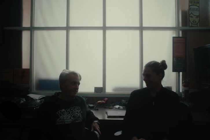 A man and a woman sitting in chairs have their backs to a white window and they look at each other